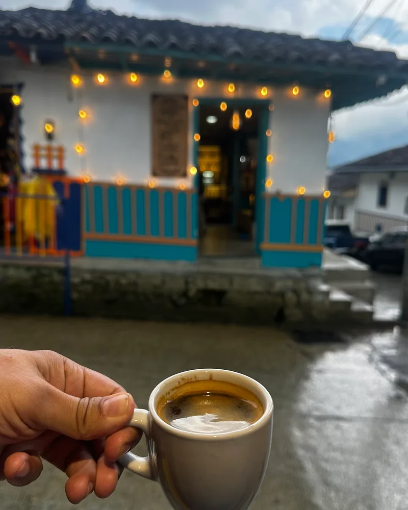 Small cup of black coffee held in front of a white building with a blue and orange wooden facade and string lights