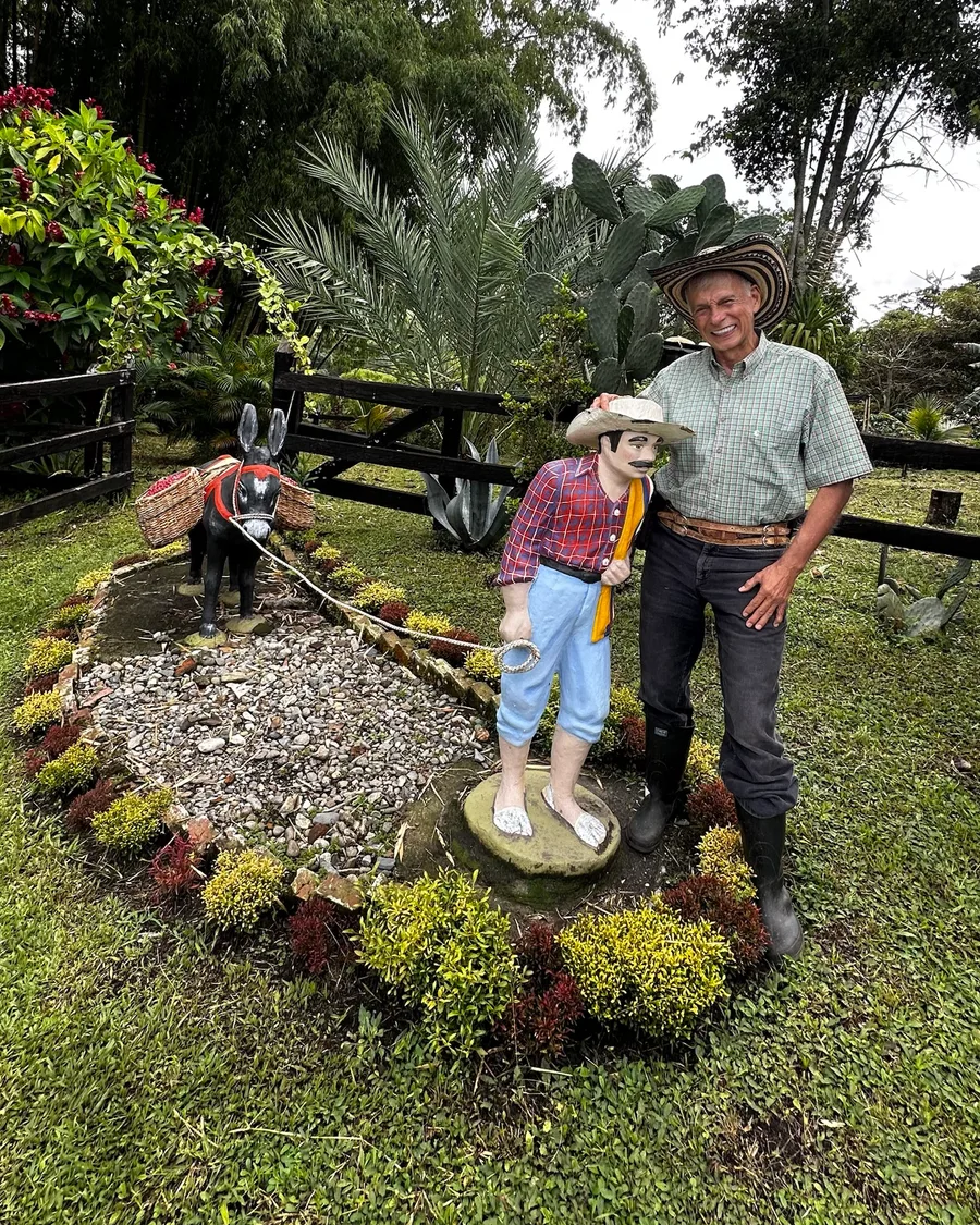 Smiling man in a sombrero and a green checked shirt standing beside a painted statue of a coffee grower leading a mule with baskets, in a garden